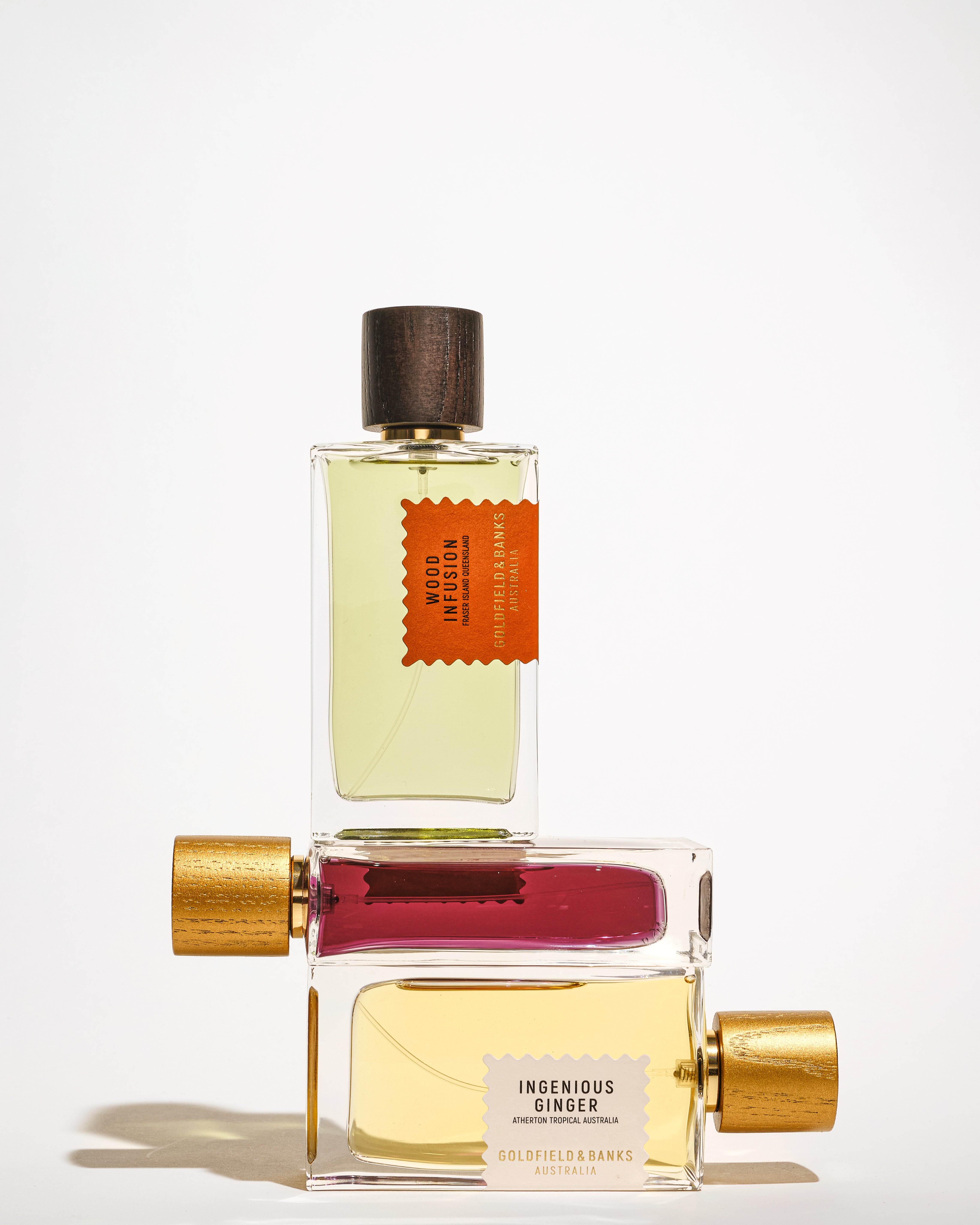Goldfield & Banks perfume bottles stacked in a minimalist composition, featuring Ingenious Ginger, Southern Bloom and Wood Infusion fragrances with natural wooden and gold caps on a clean white background.